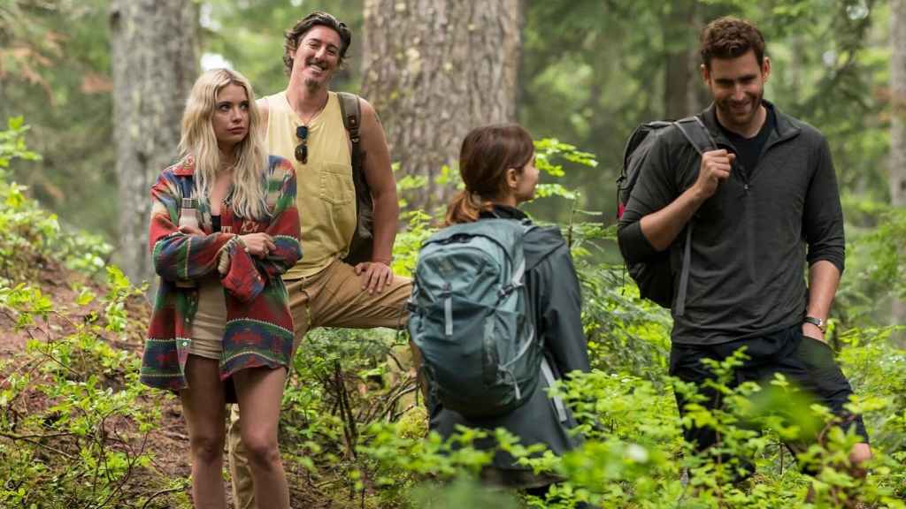 A still from the show 'Wilderness' in which four people stand in a lush forest, dressed in casual outdoor clothing, appearing to be on a hike. On the left, a blonde woman in a colorful oversized jacket and shorts stands with her arms crossed, looking displeased. Beside her, a man in a yellow tank top and beige pants leans on a tree with a cheerful smile. To the right, a woman with a ponytail and a large hiking backpack faces a man in a dark long-sleeve shirt and shorts, who is smiling and carrying a backpack. The group appears to be engaged in a conversation amid the dense greenery and tall trees.