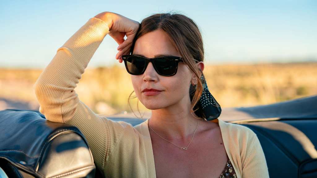 A still image from the TV series 'Wilderness' featuring Jenna Coleman as Liv Taylor, sitting in a convertible, looking in the distance, wearing sunglasses, and yellow sweater.