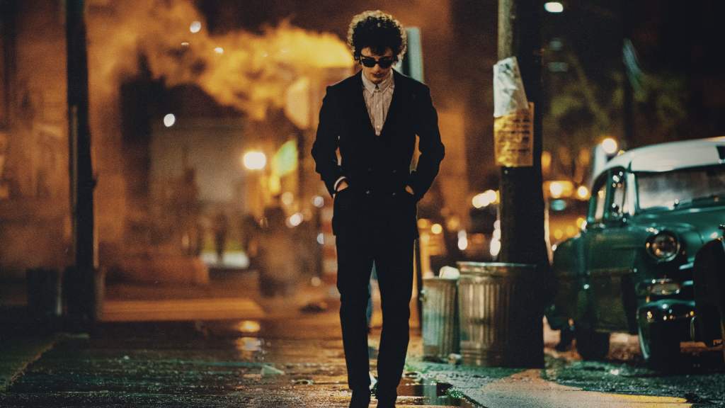 Timothée Chalamet as Bob Dylan in 'A Complete Unknown,' walking down a dimly lit street in a 1960s setting, dressed in a dark suit, with his hands in his pockets, and illuminated by the glow of streetlights and vintage cars in the background.