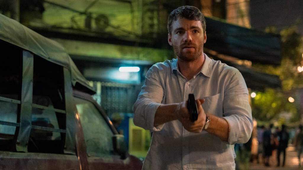 Gabriel Basso as Peter Sutherland in The Night Agent Season 2, he's wearing a white shirt and holding a gun in a street in Bangkok at night.