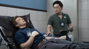 A promotional still from TV series Dexter: Original Sin featuring Patrick Gibson as young Dexter Morgan lying on a stretcher with Alex Shimizu as Vince Masuka stadning next to him.