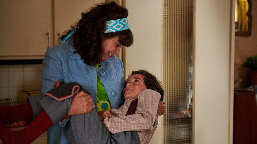 Leïla Bekhti as Esther Perez in Ma Mère, Dieu, et Sylvie Vartan or Once upon my Mother movie. She's wearing 1960s style garb and holding a small boy in her arms, looking lovingly at him.