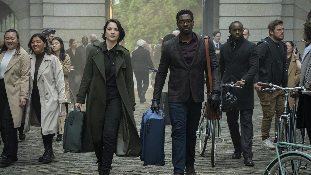 Mariana Novak as Talamasca Agent Polina Vilkov, Tongayi Chirisa as Ciprien Grieve in Mayfair Witches Season 2, both walking in a crowd, holding a small suitcase.