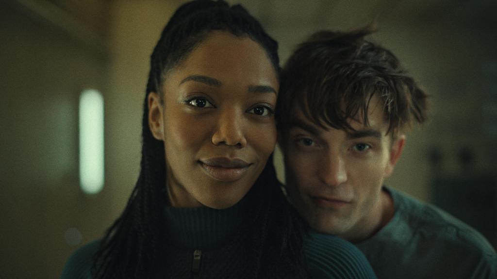 A still from the movie Mickey 17 featuring Naomie Ackie as Nasha smiling looking slightly off camera, and Robert Pattinson as Mickey Barnes, with his chin on her left shoulder looking smilingly at the camera.