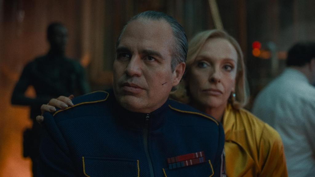 A still from the movie Mickey 17 featuring Mark Ruffalo as Kenneth Marshall, looking menacing in a military general-like outfit, behind him Toni Colette as his wife Ylfa in a gold silk blouse.