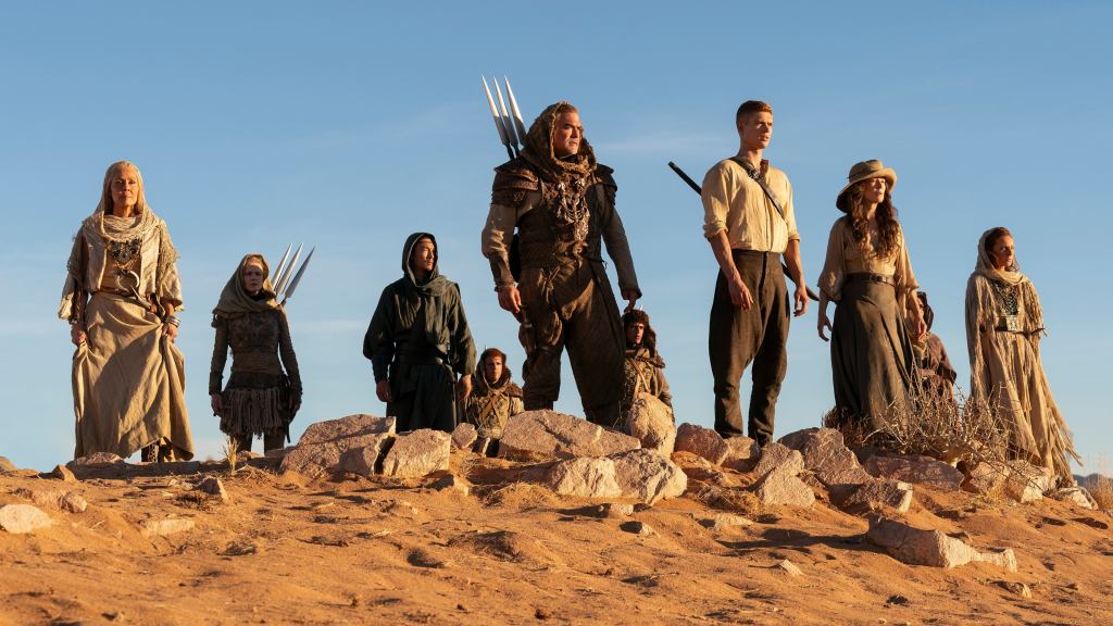 A promotional still from the third season of The Wheel of Time TV series. There's an array of people in a desertic environment looking into the distance. some of them carry spears or swords.  