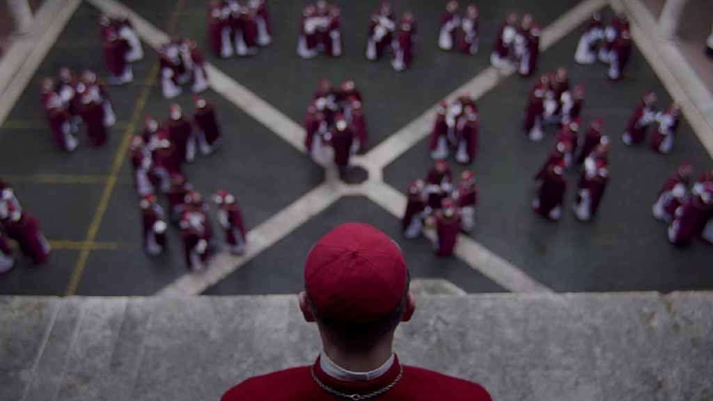 Still from the movie adaptation of Robert Harris' book Conclave, featuring an aerial shot of a courtyard with several clumps of cardinals engaged in conversation with one Cardinal in the foreground, on a high level looking down at them in the inner courtyard.
