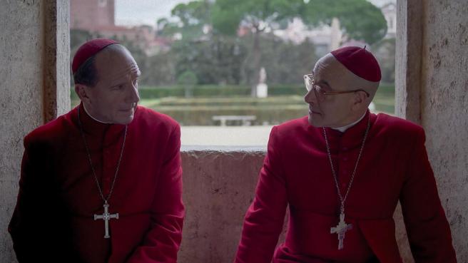 Still from the movie adaptation of Robert Harris' book Conclave, featuring Ralph Fiennes and Stanley Tucci in Cardinal garb, sitting next to each other having a deep conversation. In the background a Garden with a small pond is visible.