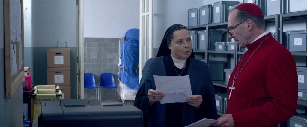 Still from the movie adaptation of Robert Harris' book Conclave, featuring Ralph Fiennes dressed in red Cardinal garb, holding some documents, talking to a none dressed in blue (Isabella Rossellini) who's also holding a piece of paper.