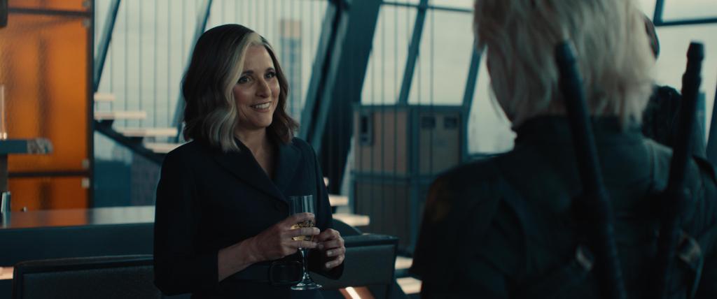 promotional still from Thunderbolts featuring Julia Louis-Dreyfus as Valentina with a smile on her face, she wears a suit and holding a champagne flute, while looking at Florence Pugh as Yelena