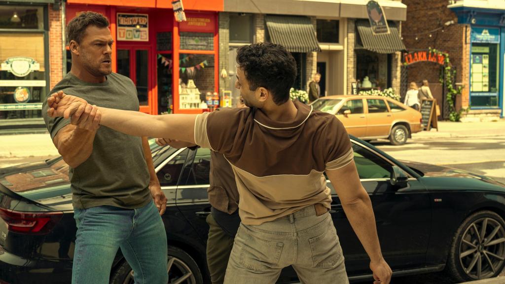 Alan Ritchson as Jack Reacher twisting a guy's arm next to a car in Reacher season 3.