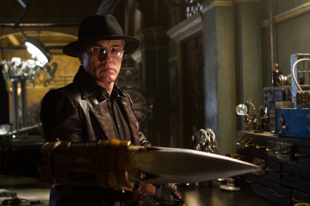 A promotional still from Alita: Battle Angel featuring Christoph Waltz wearing a cowboy hat, glasses, and a leather jacket in a mechanic lab, looking at a sharp knife
