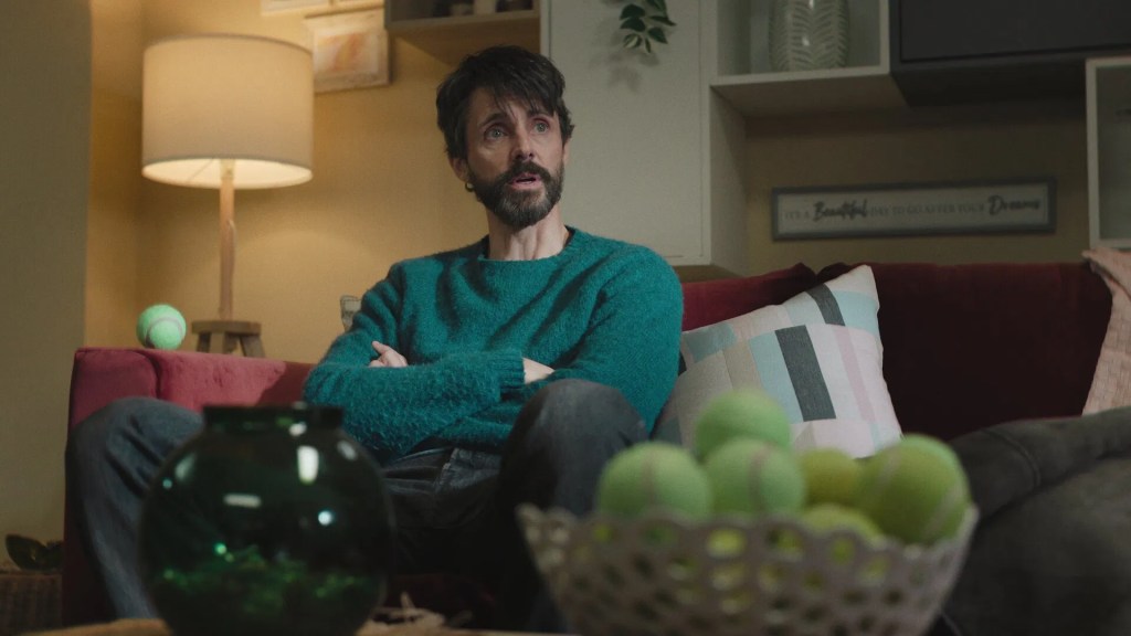 A still image from Netflix's Dept. Q season one featuring Matthew Goode as Carl Morck, sitting on a red couch in a psychologist office. He's wearing a green jumper and black jeans.