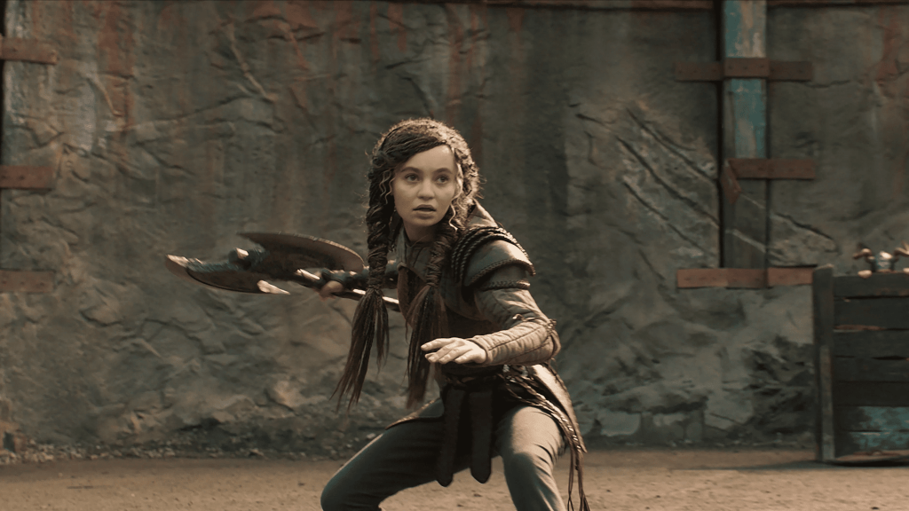 A promotional still from How to Train Your Dragon live-action movie featuring a young woman (Nico Parker as Astrid) in a battle ready stance while holding an axe.
