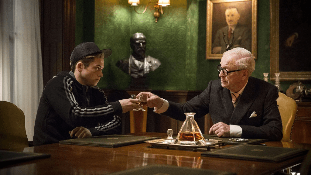 A promotional still for Kingsman: The Secret Service movie featuring Taron Egerton as Gary "Eggsy Unwin in his street clothes cheering Cognac with Michael Caine as Chester King in an expensive looking room.