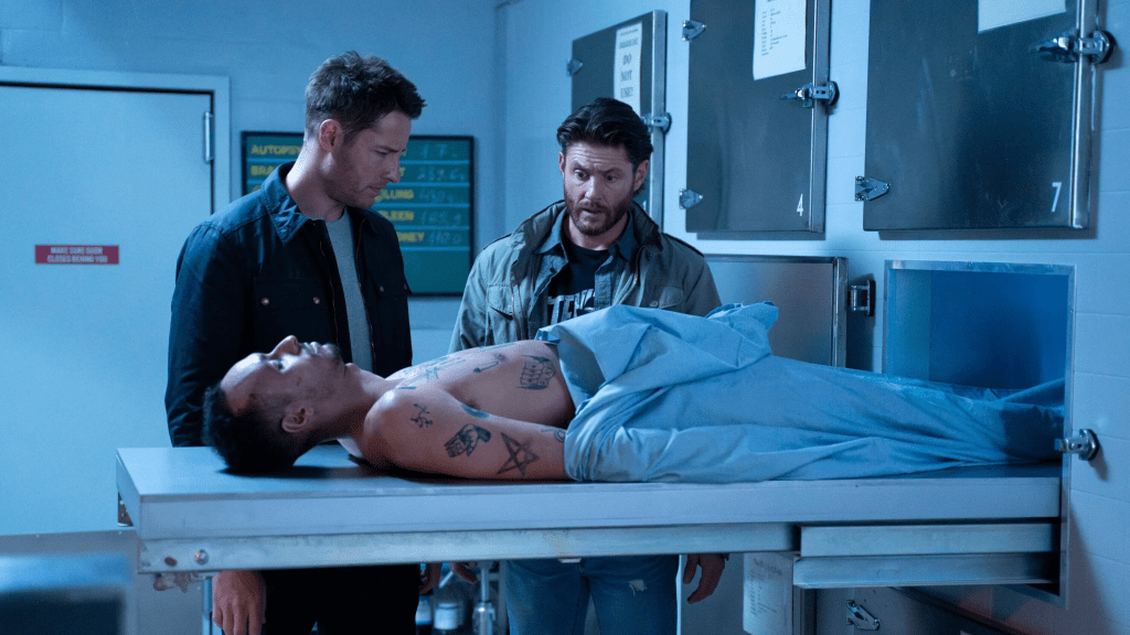 A still from TV show Tracker season one featuring two men in a morgue next to the body of man. One man (Justin Hartley as Colton Shaw) wears a dark jacket and gray t-shirt looking confused at the surprise-looking man (Jensen Ackles as Russell Shaw) next to him.