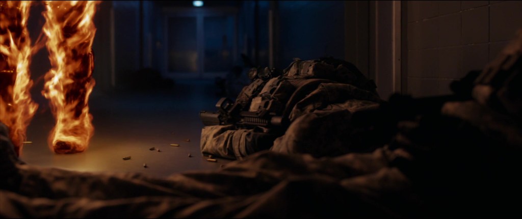 A still from 2015's Fantastic Four movie featuring a dark sinister hallway with dead soldiers in the foreground and two legs engulfed with flames walking past 