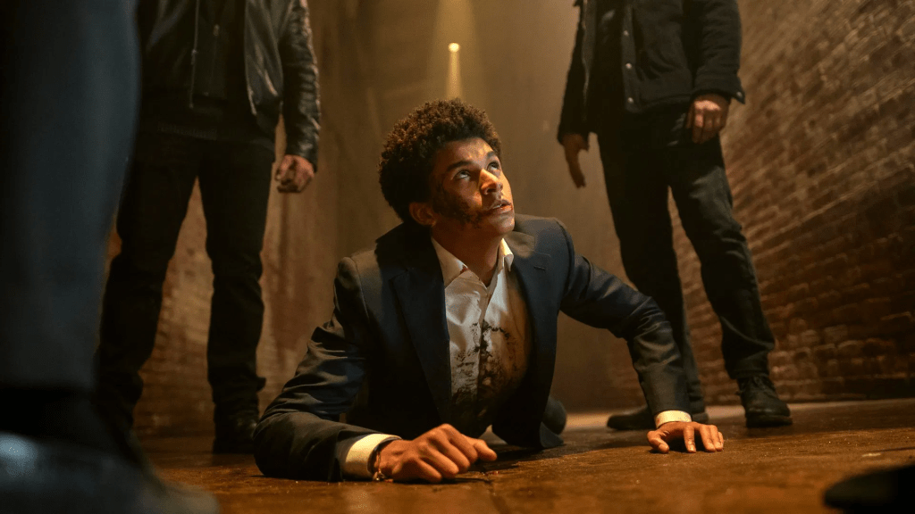 Jaden Michael as Mickey Bolitar in Harlen Coben's Shelter in a suit, bruised and with a bloodied shirt, lies on the floor of a dimly lit alleyway, looking up with a mix of fear and defiance. Three men in dark clothing stand over him, their faces mostly obscured, creating a tense and threatening atmosphere.