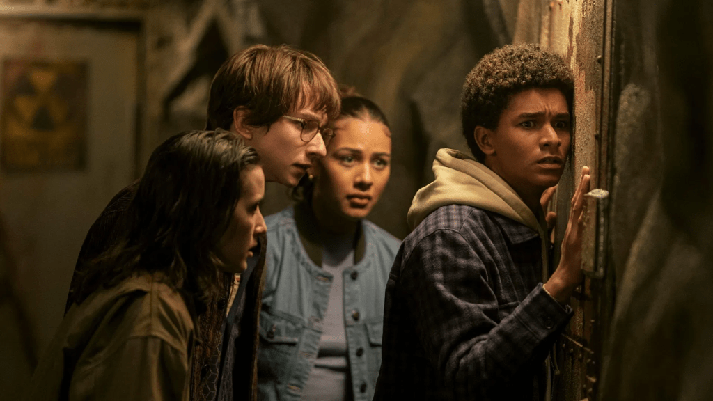 A still image from Harlan Coben's Shelter with Abby Corrigan, Adrian Greensmith, Jaden Michael, and Sage Linder stand closely together in a dimly lit, tense setting. One boy presses his ear against a rusted metal door with a worried expression, while the others look on anxiously behind him.