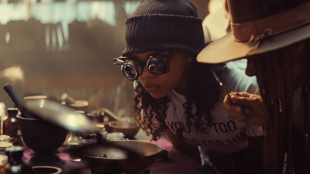 Still image from TV series Ironheart featuring a young woman (Regan Aliyah) wearing a dark beanie and futuristic steampunk-style magnifying goggles leans over a table covered with bowls, jars, and scientific tools. She is intently examining something, while another person beside her, wearing a wide-brimmed hat, holds a small spoon. Warm, dramatic lighting creates a moody, alchemical atmosphere.