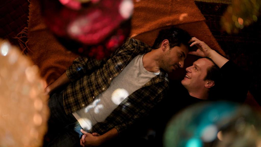Karan Soni, and Jonathan Groff in A Nice Indian Boy, both lying on a bed looking at each other.