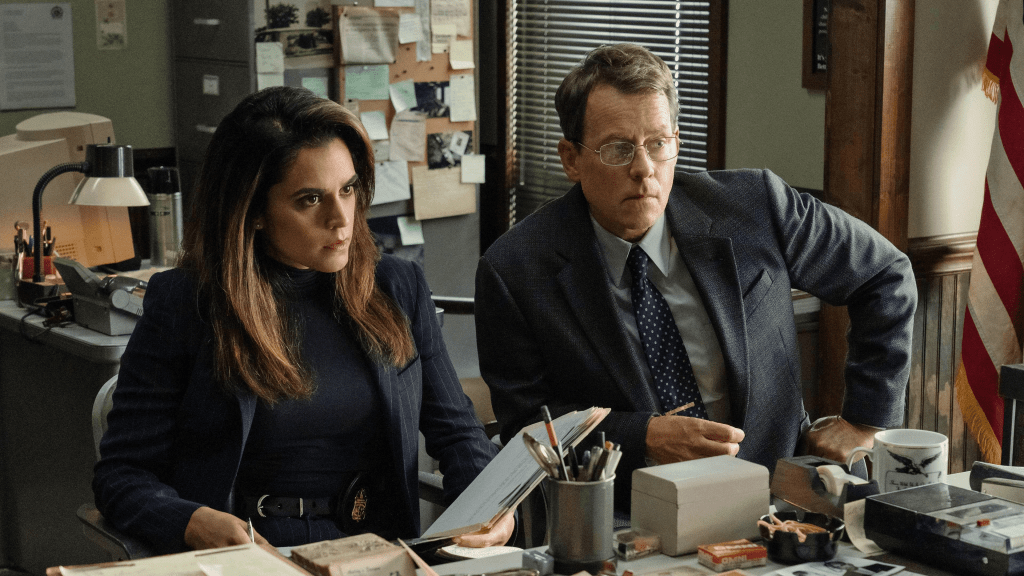 Sepideh Moafi and Greg Kinnear in Apple TV+ Black Bird, both are in an office, sat in from of a desk looking at someone off screen.