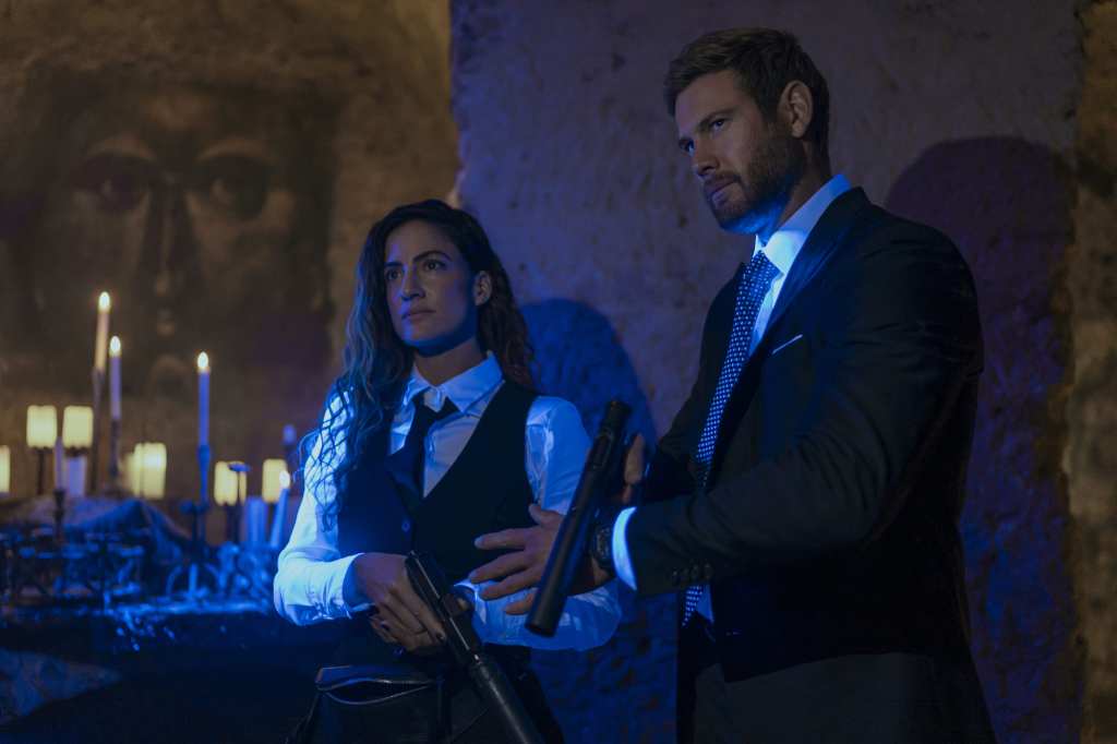 Rona-Lee Shimon and Tom Hopper in The Terminal List: Dark Wolf (2025) both holding hand guns with silencers looking away in a club setting with their white shirts glowing blue.