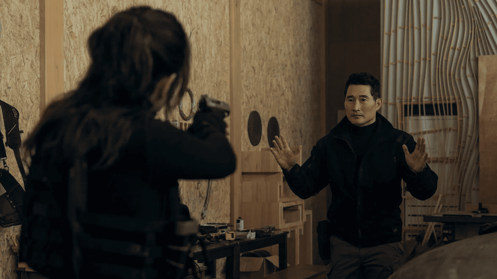 A man raises his hands in surrender as a woman aims a gun at him in a dimly lit room, a still from the tv series Butterfly.