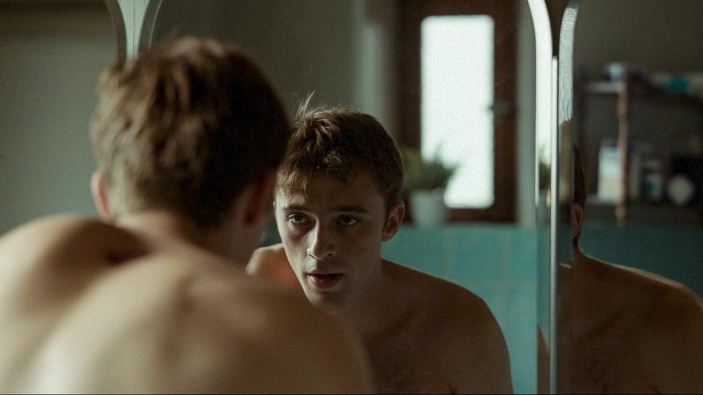 A young man looking at his reflection on a mirror, he's shirtless, and looks tired and contemplative. A scene from The Quiet Son.