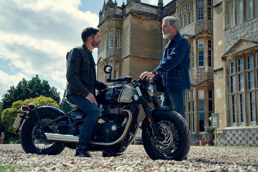 Two men talk beside a Triumph motorcycle in front of a large historic mansion, scene from The Thursday Murder Club.