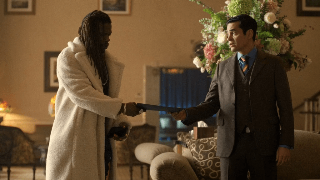 A scene from Will Trent season 3. The image shows actors Antwayn Hopper as Rafael Wexford draped in a long, white fur-like coat extending what looks like a folder or document to Ramon Rodriguez as GBI Agent Trent in a sharp, dark suit.