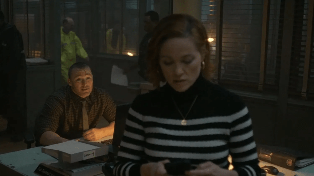 Detective Angie Polaski (Erika Christensen) on her phone with Ormewood (Jake McLaughlin) behind her at his desk in the precinct