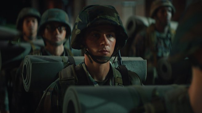 A young Marine recruit, straight-face and geared up is lining up with other recruits in the dark. A still from Boots.