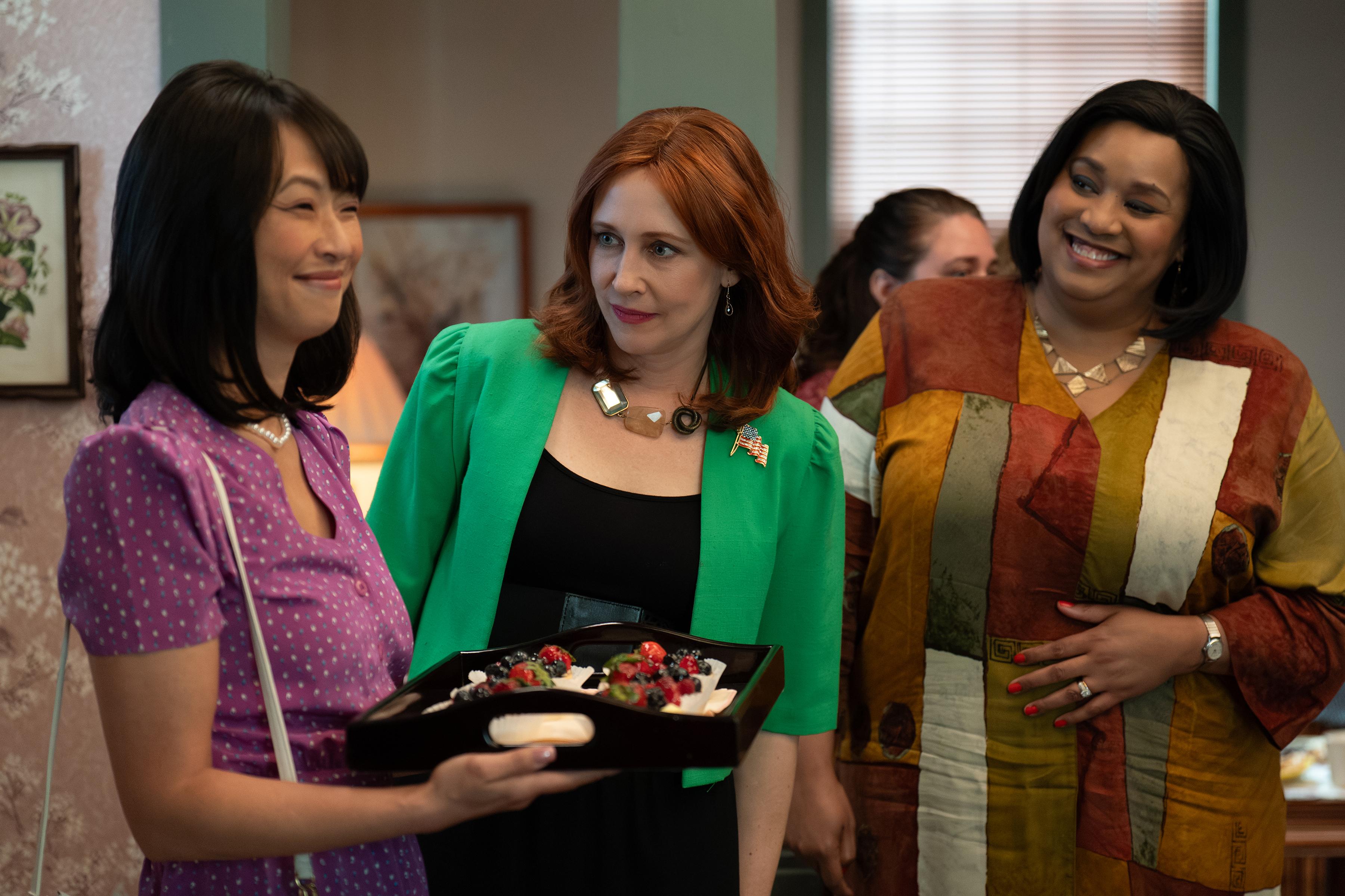 Three women share a lighthearted moment indoors, their bright clothing contrasting warmly with the muted tones of the room as one of them holds a tray of fruit-topped desserts.