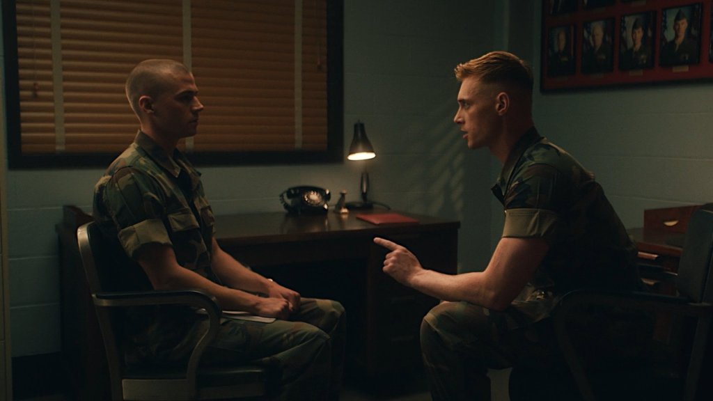 A young Marine recruit, in his green uniform, sitting back straight facing a sat drill instructor sternly pointing at him.