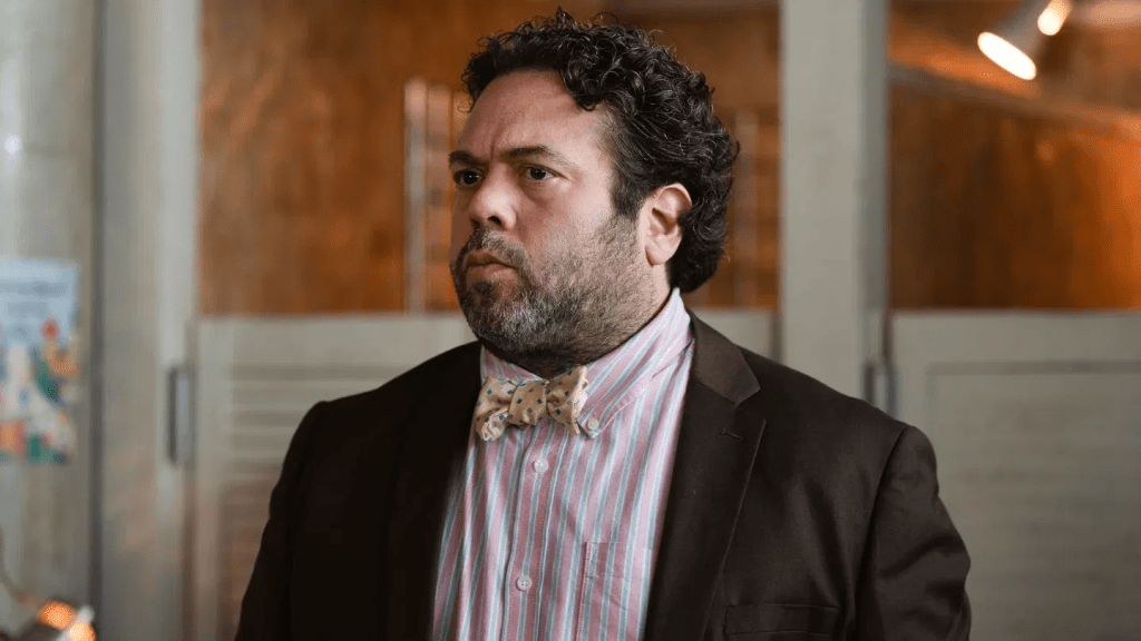 A courtroom scene featuring a man with dark curly hair and a full beard wearing a pink and blue striped shirt with a yellow bow tie and a dark brown suit jacket, looking off to the side with a serious expression.