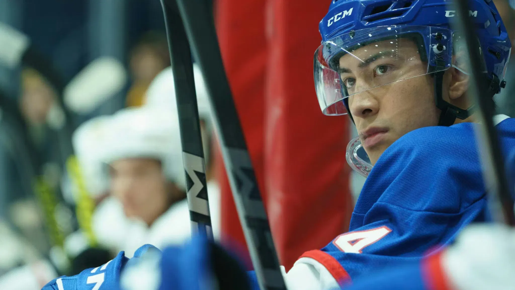 Shane Hollander (Hudson Williams) in a blue jersey with a clear visor and blue helmet, looking intently at the game.