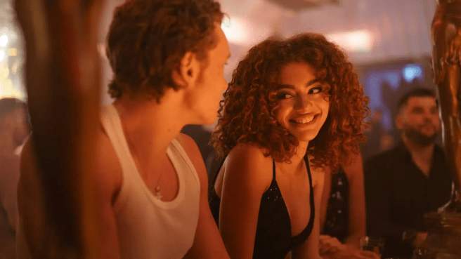 Svetlana (Ksenia Daniela Kharlamova) in a dimly lit bar smiling at Ilya (Connor Storrie) in Heated Rivalry season 1.