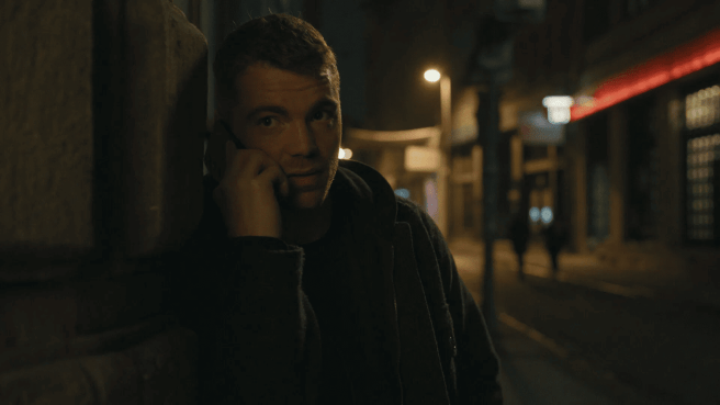 Night Agent, Peter Sullivan, leans against a wall at night holding a phone to his ear.