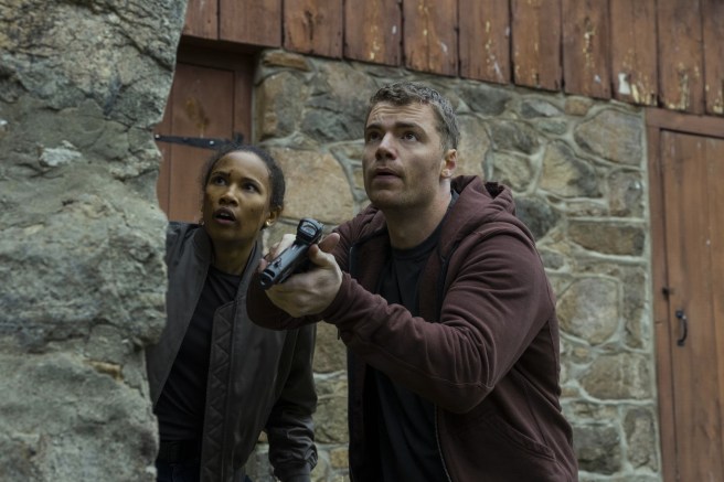 Peter Sullivan (Gabriel Basso) and Chelsea Harrington (Fola Evans-Akingbola), in The Night Agent Season 3, hiding behind a stone wall, both looking up.