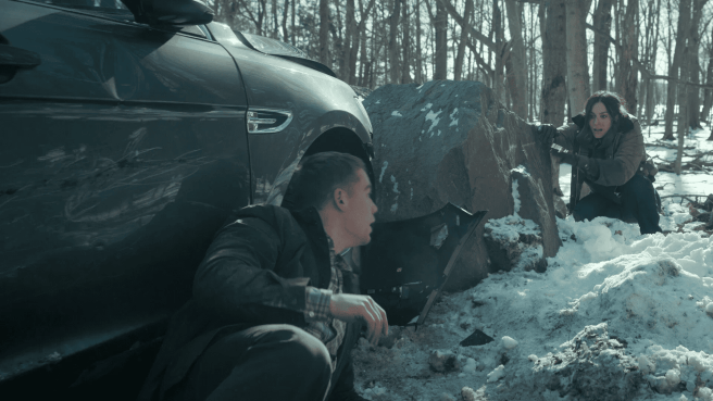 Peter Sullivan and Isabel de Leon kneeling beside a wrecked car in the snow, both looking with a concerned expression. A still from The Night Agent season 3.