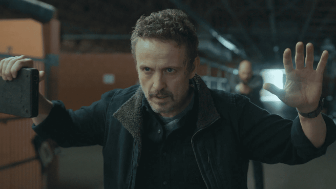 A still from The Night Agent Season 3 featuring Adam (David Lyon) in a stable holding his hands up, behind him a suited man pointing a gun.