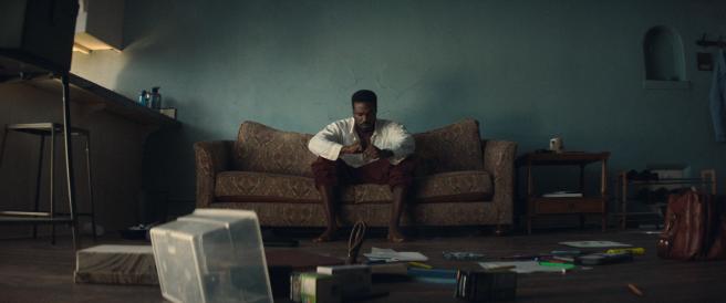 A man in a white shirt and red pants sits slumped forward on a patterned sofa in a dimly lit room, with his fists clenched.