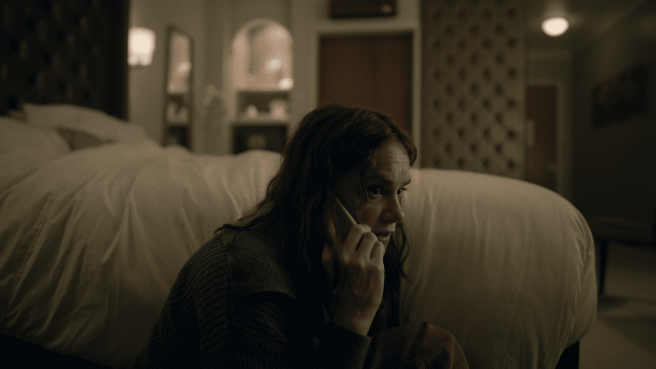 A woman sits by a large, unmade bed in a dimly lit room, speaking on a mobile phone with a look of concern.