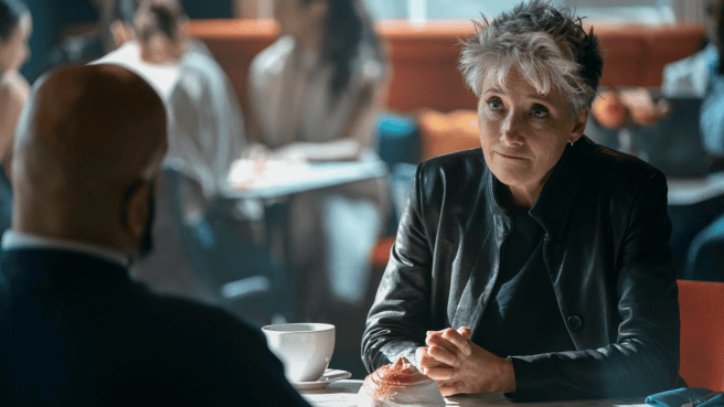 Emma Thompson as Zoe Boehm with short, spiky gray hair, wearing a black leather jacket and black top, looks intently at a man across a table in a cafe setting.