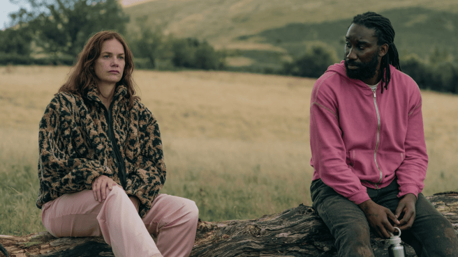 Ruth Wilson as Sarah Trafford and Nathan Stewart-Jarrett as Downey siting apart on a fallen log in a grassy, rolling landscape.