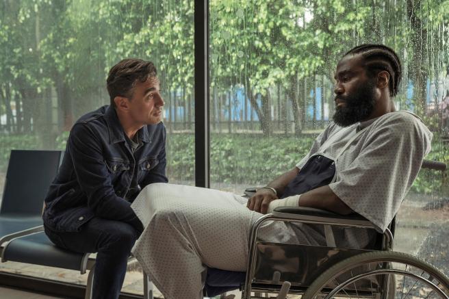 Yahya Abdul-Mateen II as John Creasy in a wheelchair wearing a hospital gown with bandages on his arm talking to Bobby Cannavale as Paul Rayburn sitting beside him.