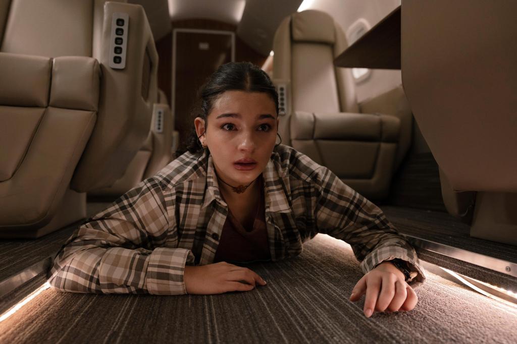 Billie Boulet as Poe Rayburn with a distressed expression is crawling on the floor of a private jet, surrounded by rows of plush leather seats.