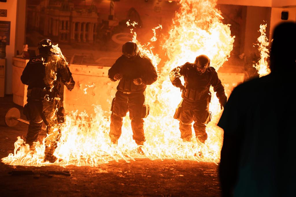 Three figures in riot gear appear to jump or move through a large wall of intense orange flames.