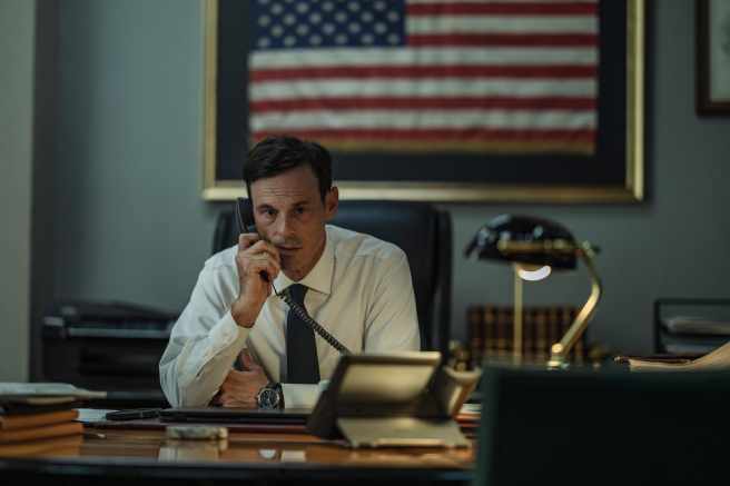Scoot McNairy in a white dress shirt and dark tie speaks on a black landline telephone while sitting at a large wooden desk, in 'Man on Fire'
